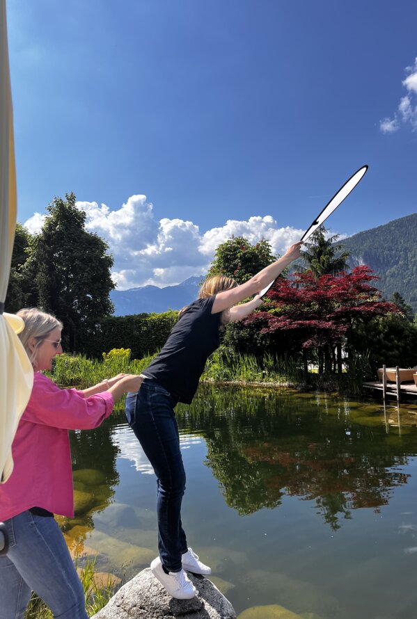 Frau beugt sich über Wasser, gehalten von anderer Frau, Bergkulisse im Hintergrund