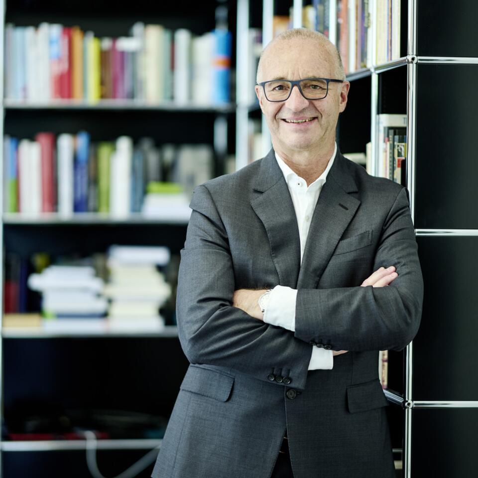 Lachender Mann mit Brille, Anzug und verschränkten Armen vor Bücherregal