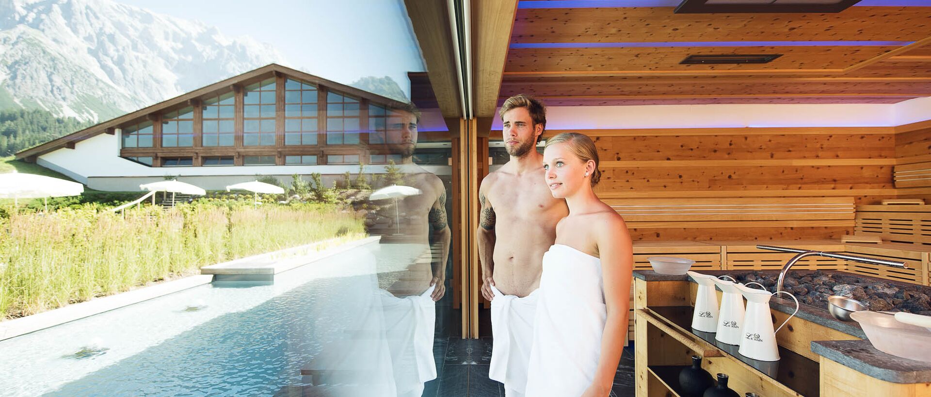 Paar in Handtüchern vor Fenster mit Pool und Bergpanorama im Wellnessbereich