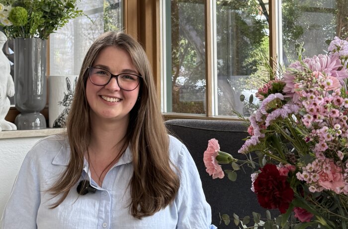Porträt einer lächelnden Frau mit Brille in einem lichtdurchfluteten Innenraum mit Blumenarrangements.