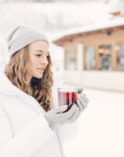 Eine Person in Winterkleidung hält draußen im Schnee ein warmes Getränk in den Händen, vor einem Gebäude mit winterlicher Umgebung im Hintergrund.