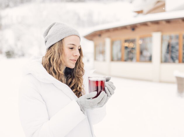 Eine Person in Winterkleidung hält draußen im Schnee ein warmes Getränk in den Händen, vor einem Gebäude mit winterlicher Umgebung im Hintergrund.
