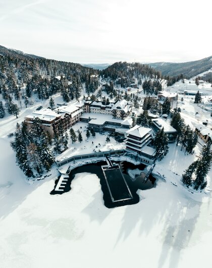 Ein Hotelresort liegt mitten in einer verschneiten Winterlandschaft mit Wäldern und einem gefrorenen See.