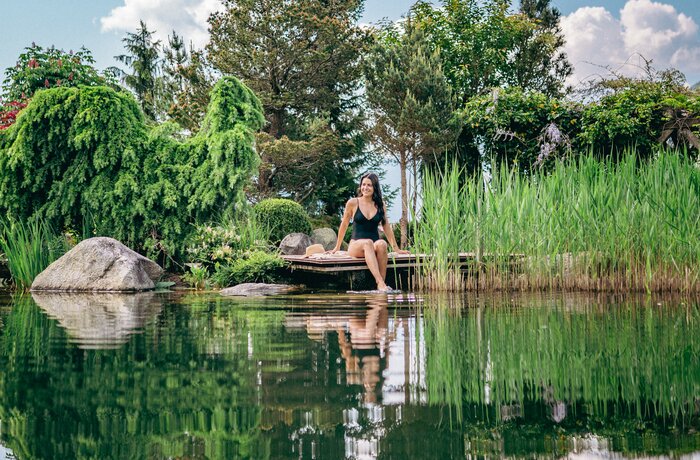 Woman sitting barefoot on wooden jetty by pond, surrounded by lush vegetation and reflections in the water