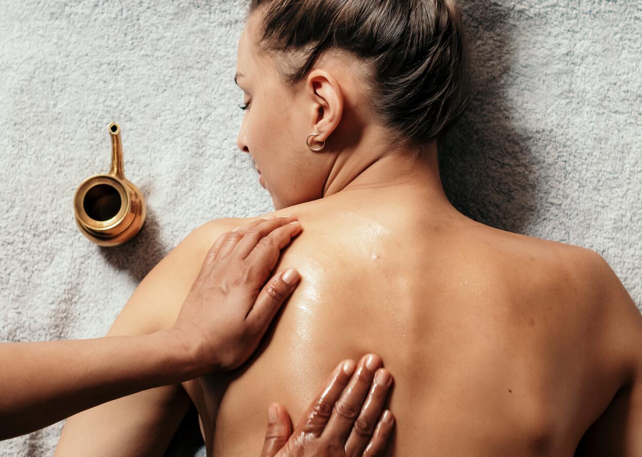 Frau liegt auf Bauch, wird mit Öl massiert, goldene Kanne daneben