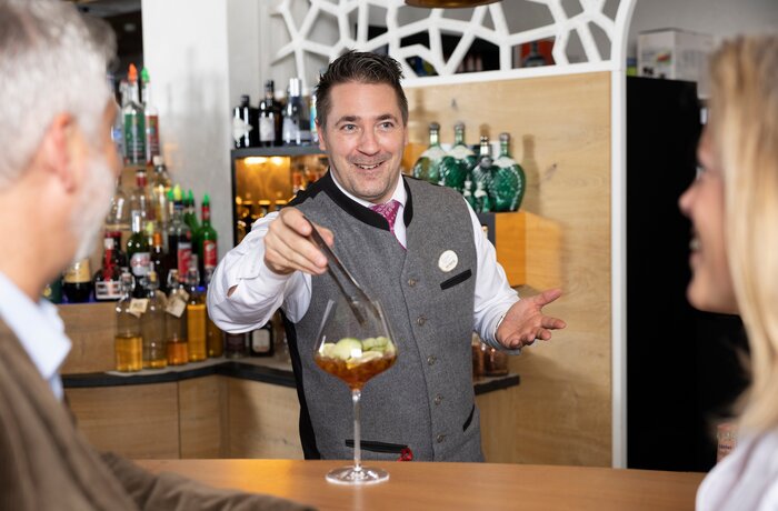 Barkeeper mixt Cocktail hinter Bar im Alpbacherhof, Pärchen im Hintergrund