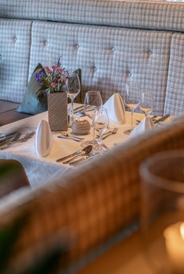 Gedeckter Restauranttisch mit weißer Tischdecke, Gläsern, Besteck und Blumenvase vor gepolsterter Sitzbank