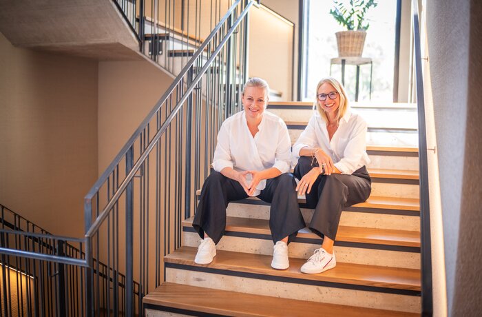 Zwei Frauen sitzen entspannt auf einer modernen Treppe und lächeln in die Kamera – stilvoll, freundlich und authentisch.