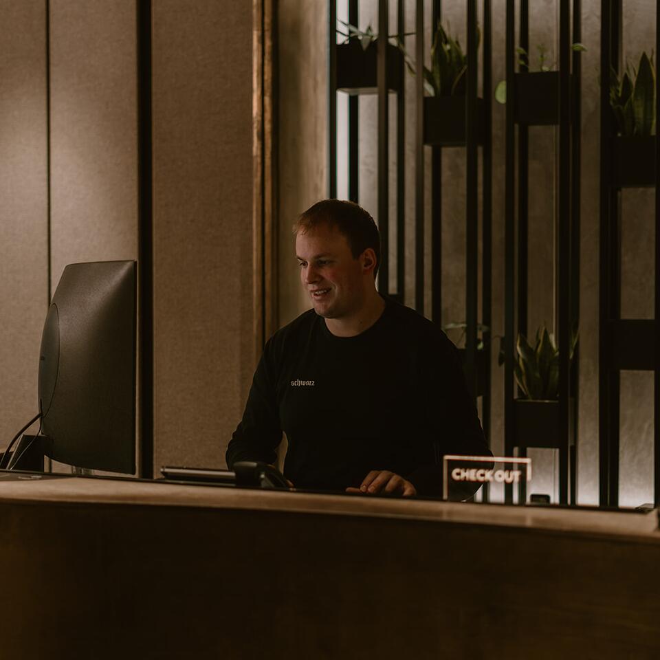 Mann in schwarzem Pullover sitzt lächelnd an einem Schreibtisch mit Computer und "Checkout"-Schild