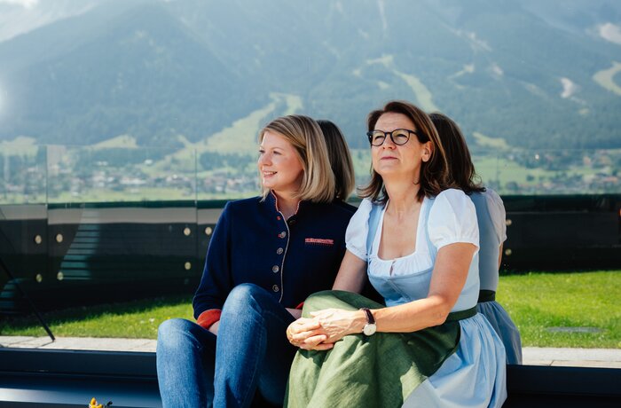Zwei Frauen sitzen draußen vor einer beeindruckenden Berglandschaft und genießen die Aussicht bei sonnigem Wetter.