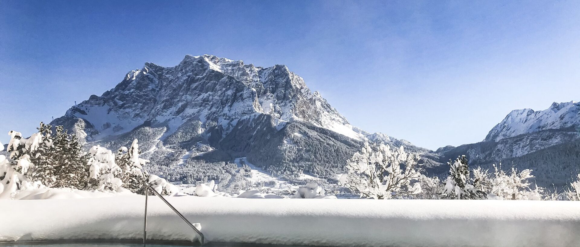 Winterlicher Pool mit Blick auf die schneebedeckte Zugspitze unter blauem Himmel