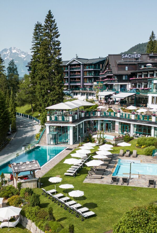 Luftaufnahme eines Wellnesshotels mit Außenpools, Liegewiesen und Gartenanlage, umgeben von hohen Bäumen und Bergen.
