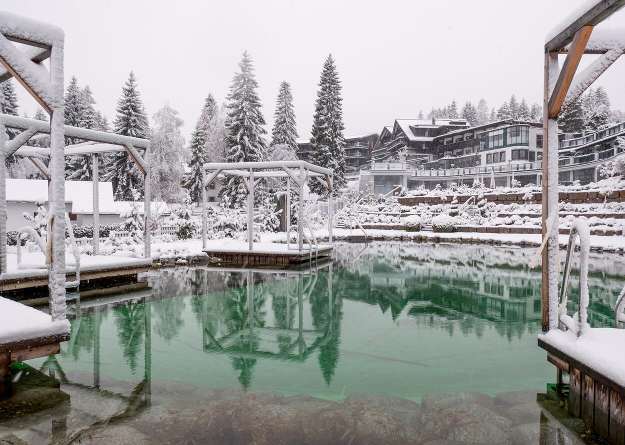 Winterlicher Naturbadesee mit schneebedeckten Bäumen und Gebäuden, Spiegelung im klaren, grünlichen Wasser
