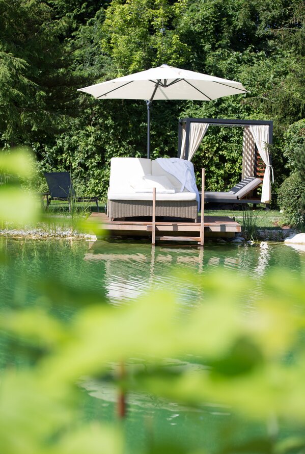 Eine gemütliche Lounge mit Sonnenschirm steht direkt an einem ruhigen Naturpool im Grünen.