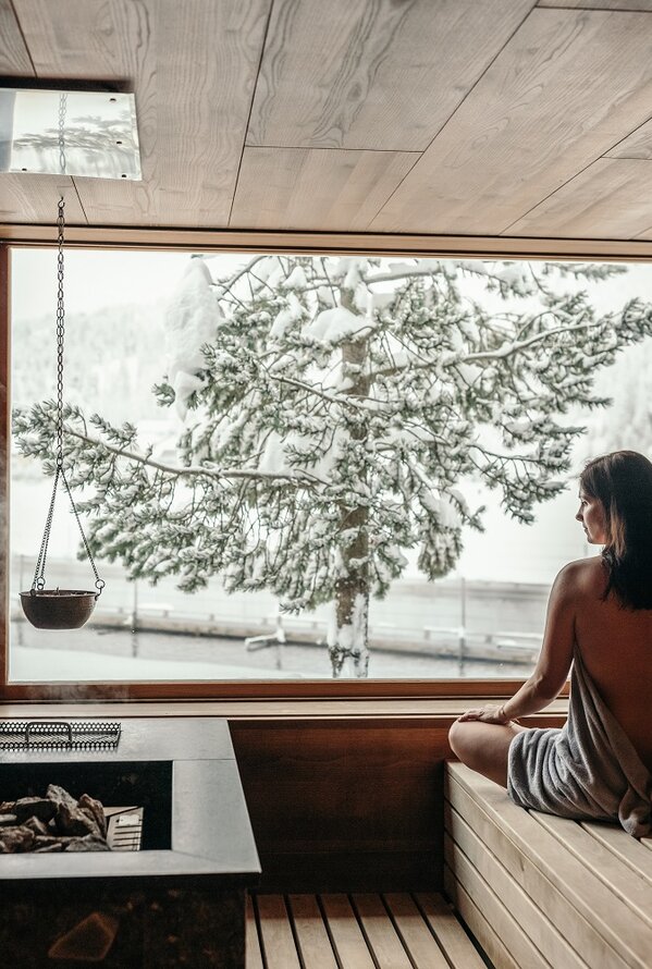 Frau in Kräutersauna blickt durch Fenster auf verschneite Landschaft