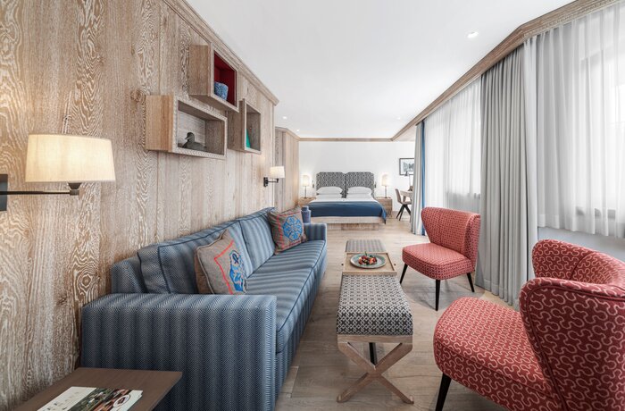 Ein modernes Hotelzimmer mit Sofa, Sesseln und einem Bett im Hintergrund. Große Fenster sorgen für viel Tageslicht.