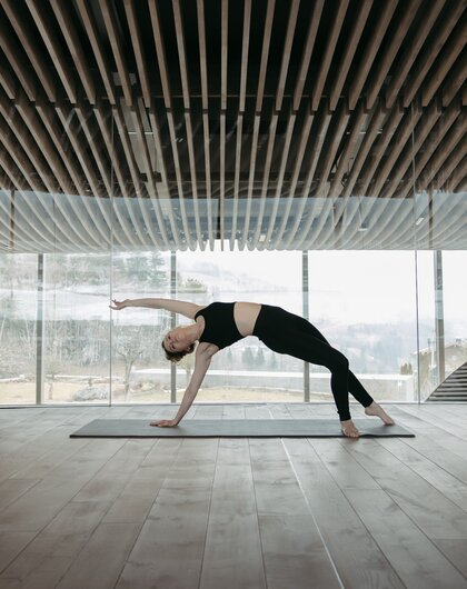 Frau macht Yoga in hellem Raum mit Holzdecke und Panoramafenster