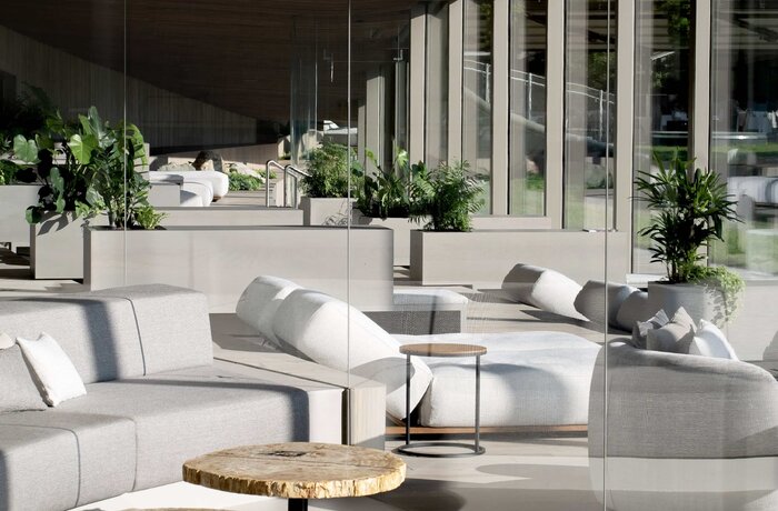 Heller, moderner Innenraum mit grauen Sofas, Holztischen und Pflanzen vor einer Glaswand