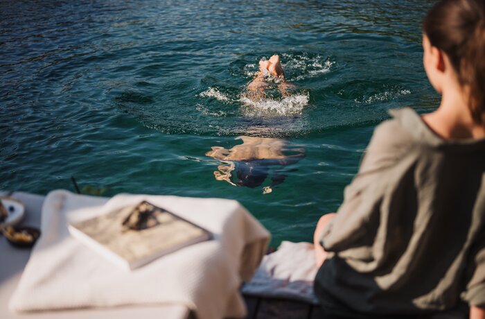 Person schwimmt im Wasser, während jemand am Ufer sitzt.