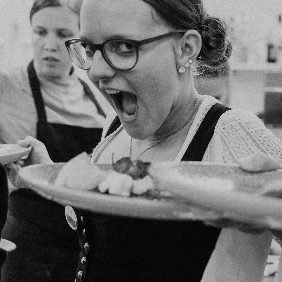 Kellnerin mit Brille und Dirndl trägt Teller mit Speisen und blickt überrascht in die Kamera