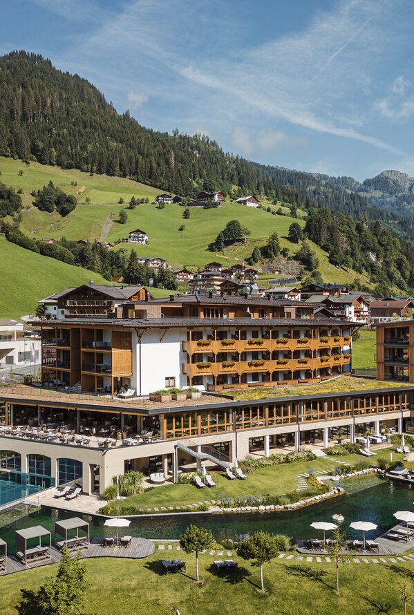 Panoramaaufnahme eines Wellnesshotels mit Infinity-Pool, Naturbadeteich und Berglandschaft