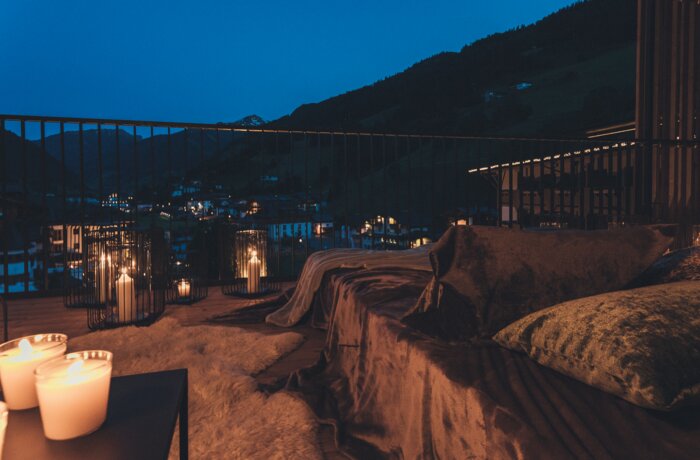 Eine gemütliche Terrasse bei Nacht mit Kerzenlicht, weichen Kissen und Blick auf ein beleuchtetes Bergdorf. Die warme Beleuchtung schafft eine entspannte und romantische Atmosphäre.