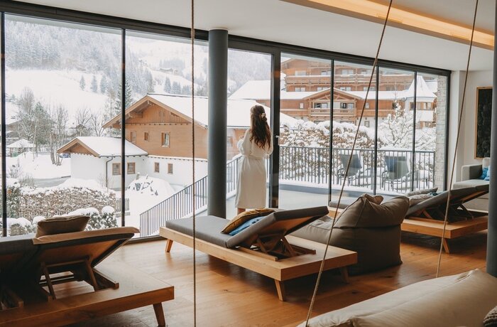 Frau im Bademantel blickt aus Ruheraumfenster auf verschneite Winterlandschaft