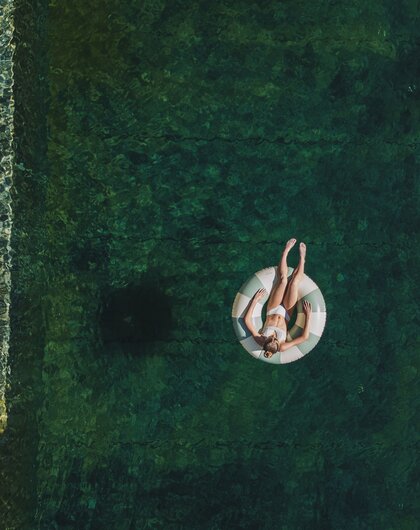 Drohnenaufnahme einer Frau auf einem Schwimmring in einem Naturpool mit klarem Wasser
