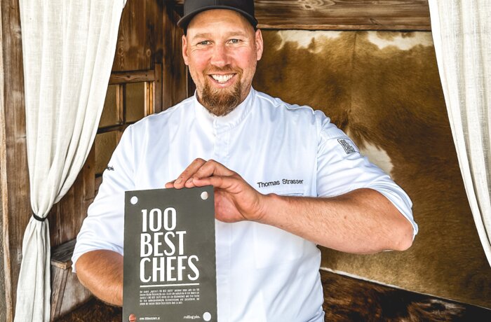 Koch Thomas Strasser mit "100 Best Chefs"-Buch vor rustikalem Hintergrund