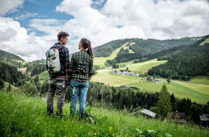 Wanderpaar blickt auf grüne Almwiese mit Häusern und bewaldeten Hügeln unter bewölktem Himmel