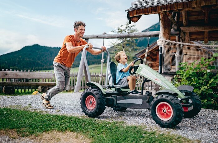 Mann schiebt lachenden Jungen in Go-Kart vor Holzhütte und Bergkulisse