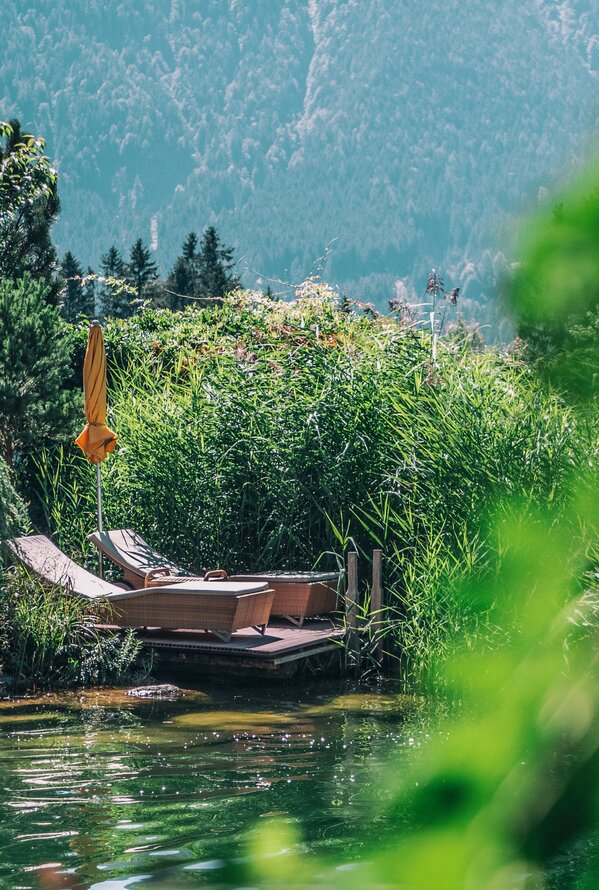 Zwei Liegestühle am Ufer eines Sees, umgeben von üppigem Grün und Bergen im Hintergrund
