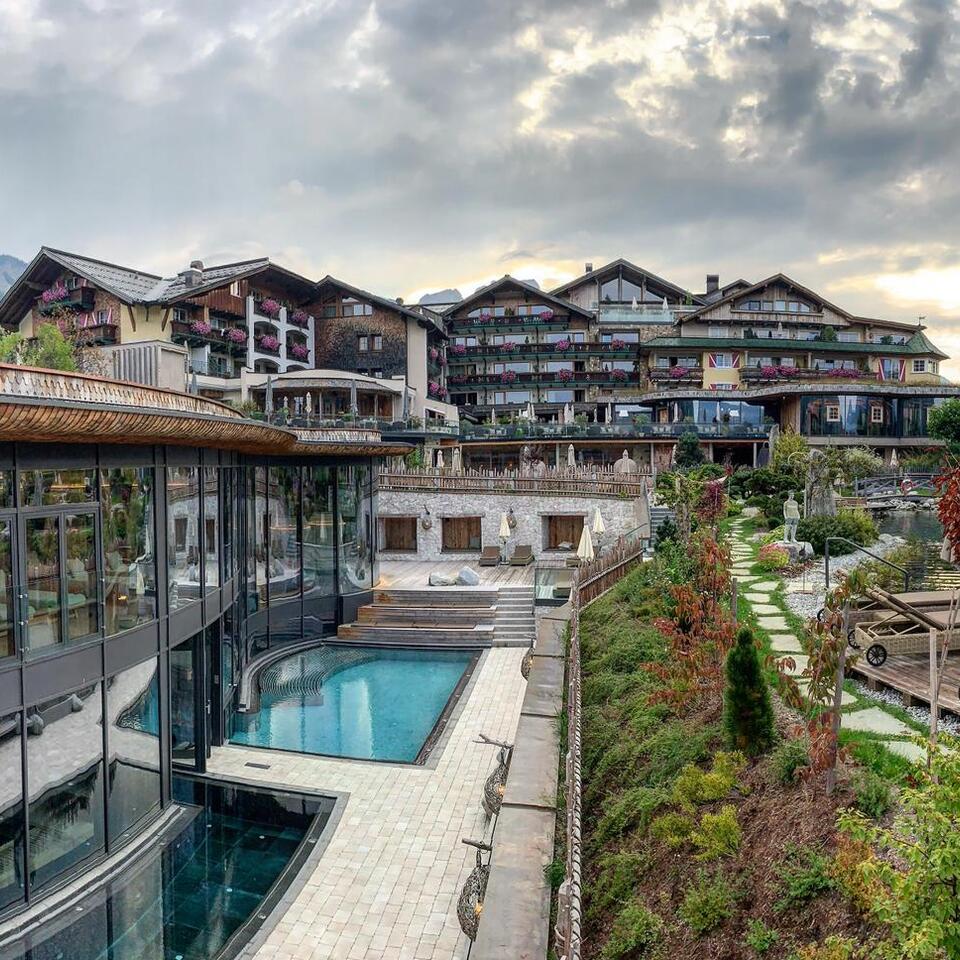 Außenansicht eines Hotels mit Pool und Gartenanlage vor bewaldeten Bergen unter bewölktem Himmel