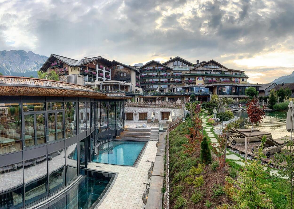 Außenansicht eines Hotels mit Pool und Gartenanlage vor bewaldeten Bergen unter bewölktem Himmel