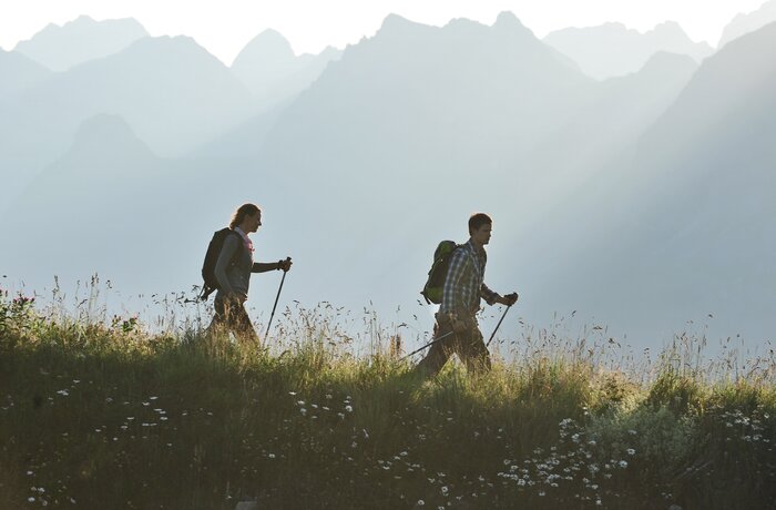 Zwei Wanderer mit Rucksäcken und Stöcken auf einem grasbewachsenen Hügel vor verschwommenen Bergen