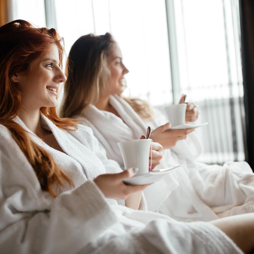 Zwei Frauen im Bademantel mit Tassen sitzen entspannt vor einem Fenster