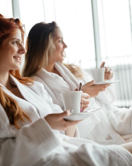 Zwei Frauen im Bademantel mit Tassen sitzen entspannt vor einem Fenster